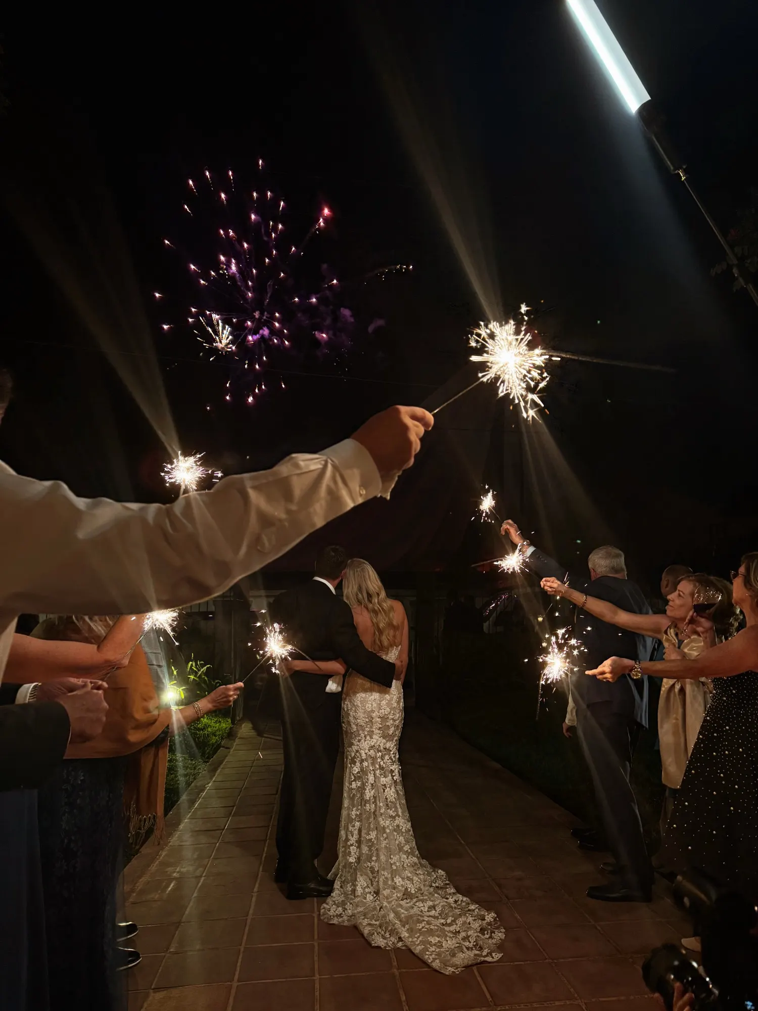 A bride and groom walking away at night through a sparkler send-off, with fireworks overhead